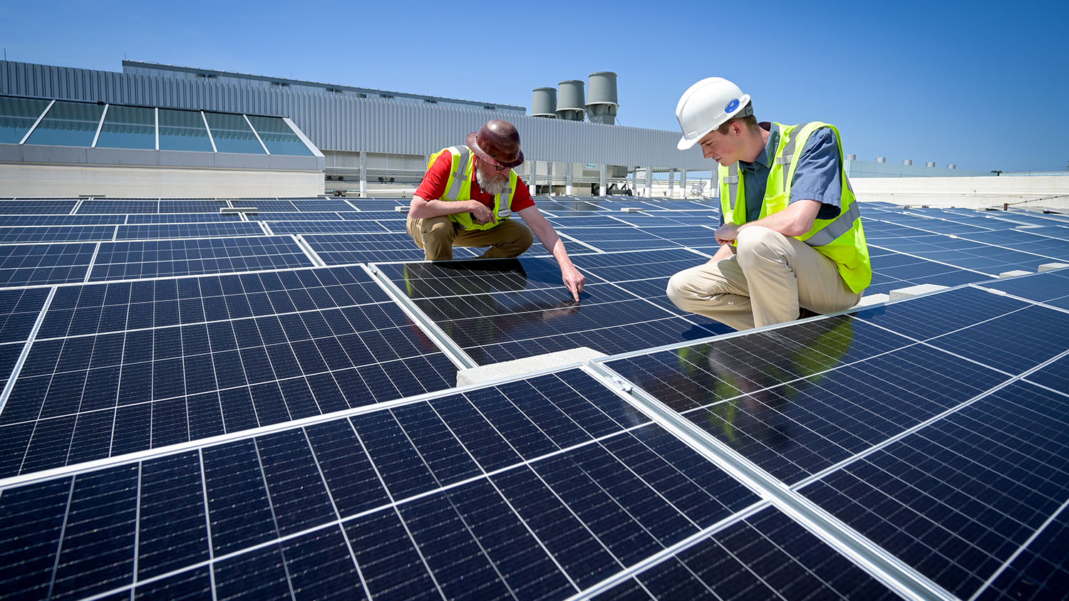 A student intern and a construction manager inspect solar panels installed on the Fitts-Woolard Engineering Building on Centennial Campus. A clear blue sky shows behind them, above the building they are working atop.