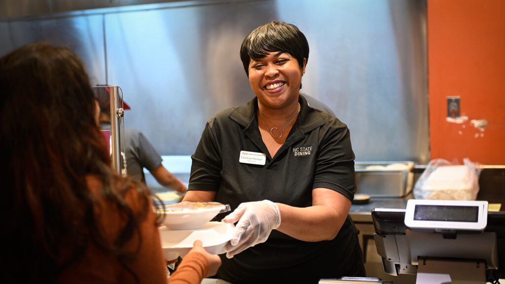 A dining employee smiles and hands food to a student.