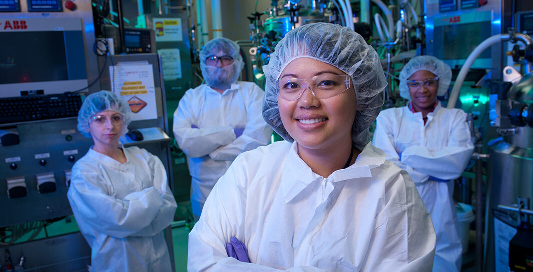 Four researchers wearing hairnets, goggles, gloves and white jumpsuits stand in a group in a biomanufacturing lab with machinery behind them.